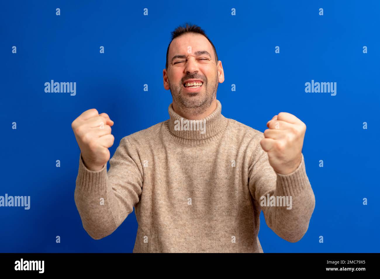 Fröhlicher, aufgeregter Hispanic mit Rollkragenpullover, isoliert auf blauem Hintergrund stehend, um den Erfolg mit erhobenen Fäusten und geschlossenen Augen zu feiern Stockfoto