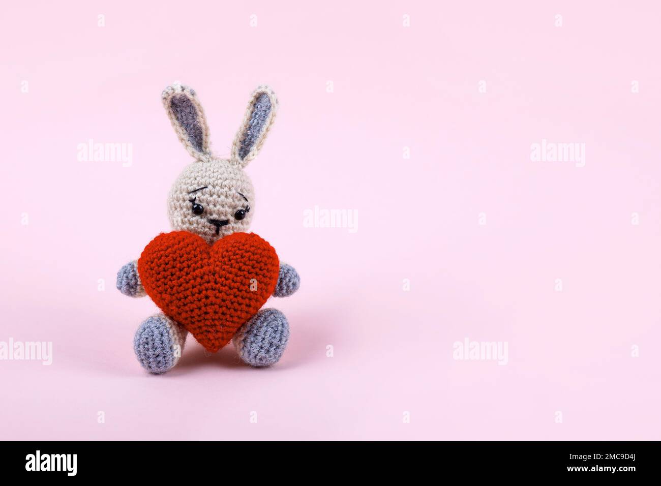 Gestricktes graues Häschen mit einem gestrickten roten Herz auf pinkfarbenem Hintergrund. Grußkarte zum Valentinstag, Muttertag und Geburtstag. Stockfoto