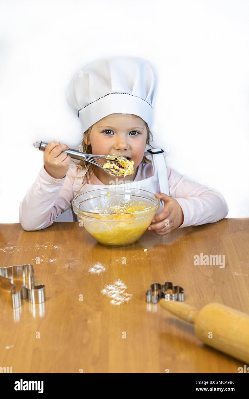 Ein kleines Mädchen kocht mit Schürze und Mütze, hält einen Schneebesen mit Teig, eine Tasse. Stockfoto