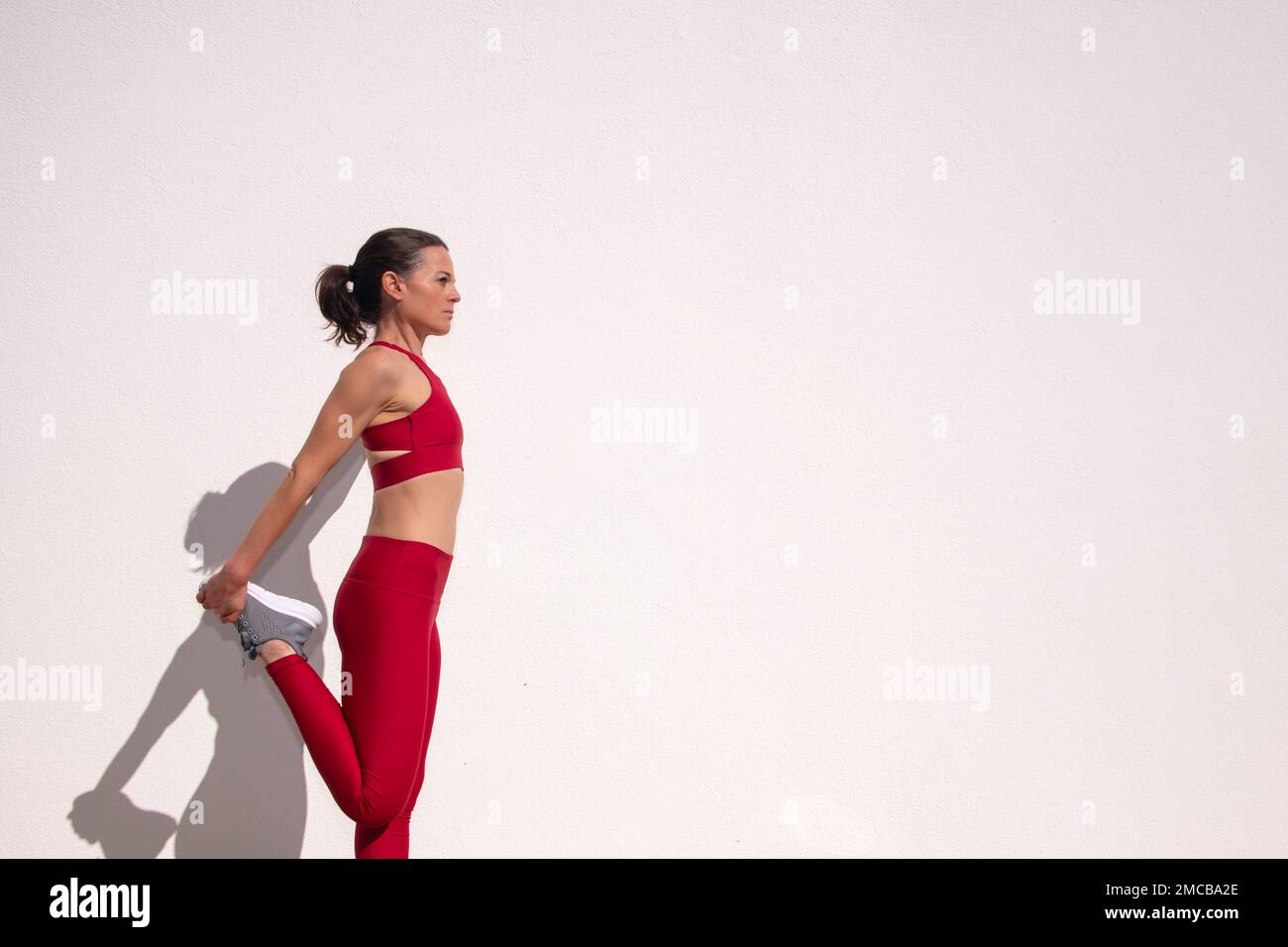 Sportliche Frau mit einem vierbeinigen Bein dehnt sich gegen eine weiße Wand in der Sonne. Stockfoto