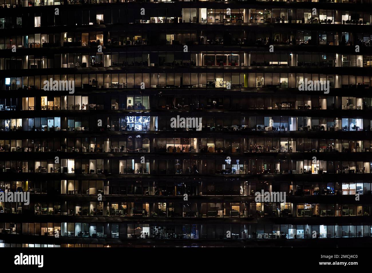 Von der Vorderseite aus blickt man auf die Nachtfassade des Gebäudes mit vielen Fenstern und Licht. Leben in jedem Fenster. Nachtsicht auf die Glasfassade eines Bürohochhauses Stockfoto
