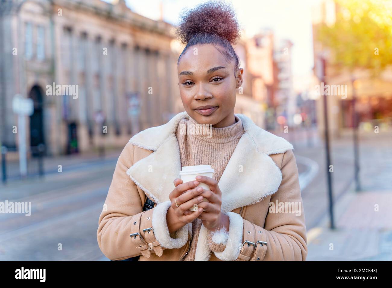 Fröhliche Afrikanerin hält eine Tasse Kaffee. Eine lächelnde braune Dame in braunem Pullover, die auf eine Straßenbahn wartet. Stockfoto