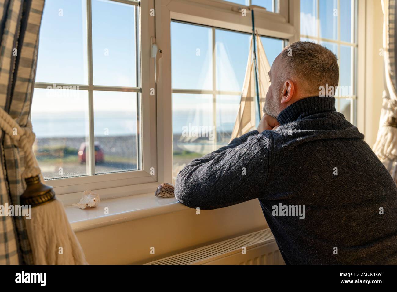 Mann im Pullover sitzt am Fenster in einem klassischen, alten Hotelzimmer mit Meerblick Stockfoto