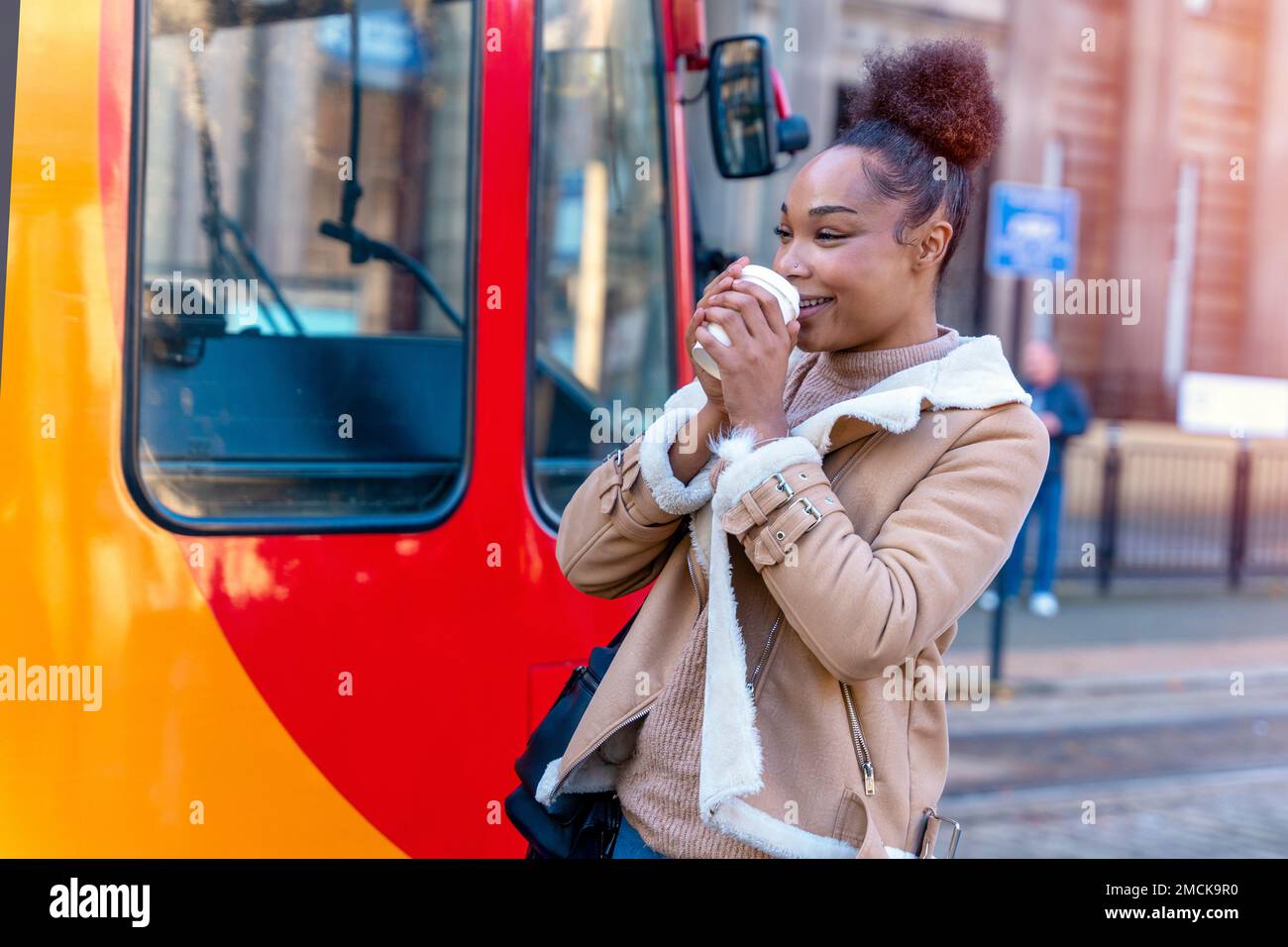 Fröhliche Afrikanerin hält eine Tasse Kaffee. Eine lächelnde braune Dame in braunem Pullover, die auf eine Straßenbahn wartet. Stockfoto