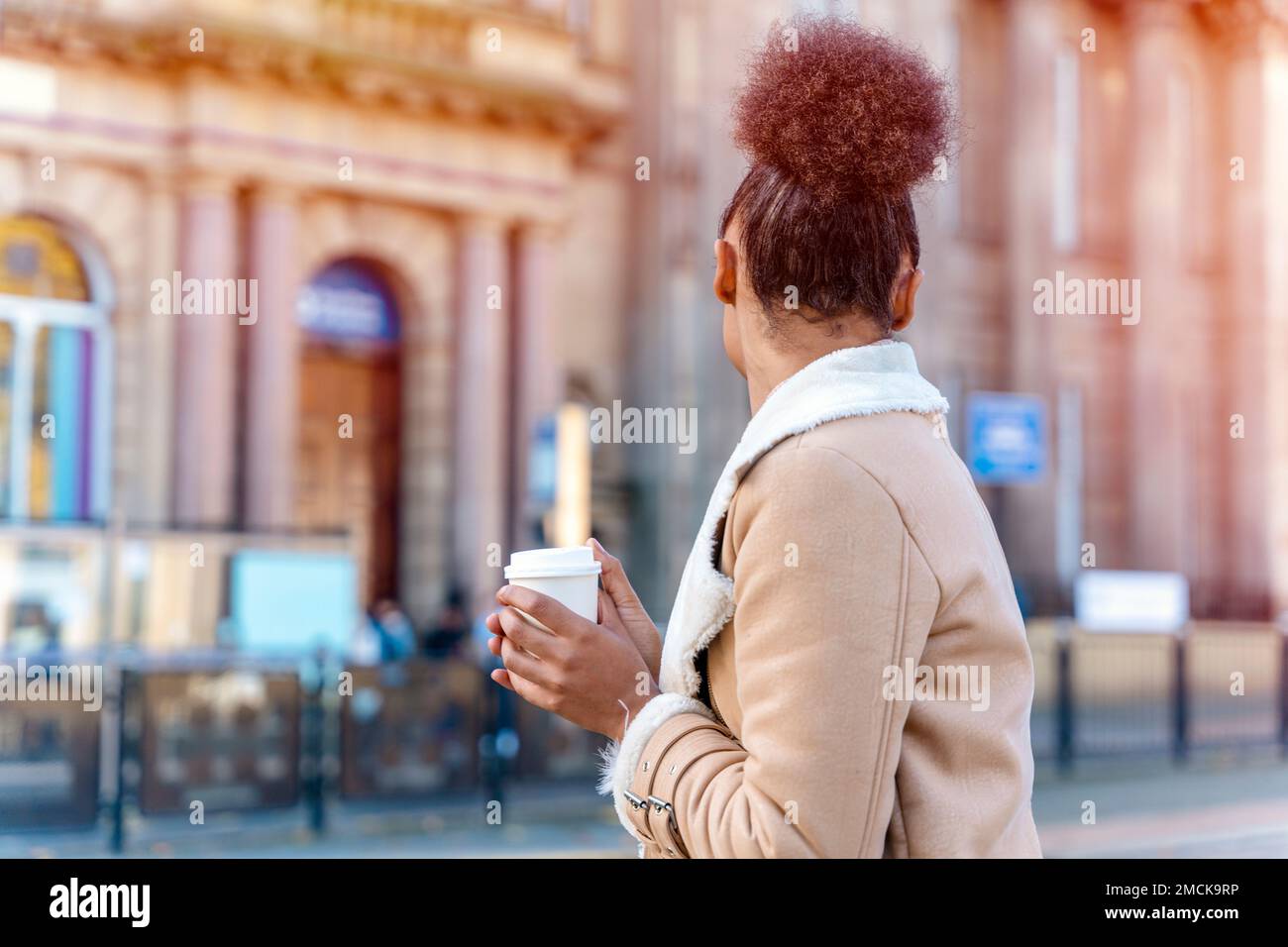 Fröhliche Afrikanerin hält eine Tasse Kaffee. Eine lächelnde braune Dame in braunem Pullover, die auf eine Straßenbahn wartet. Stockfoto