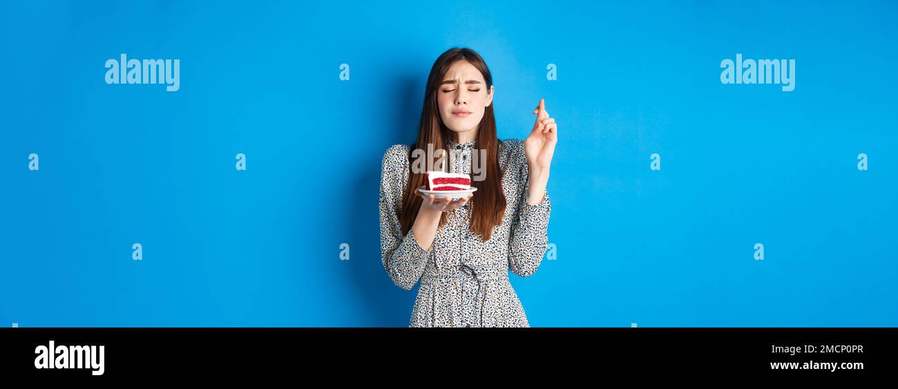 Hoffnungsvolles Geburtstagskind Wunsch für Traum wahr werden, Kreuzfinger mit geschlossenen Augen, halten Kuchen mit Kerze, blauer Hintergrund Stockfoto