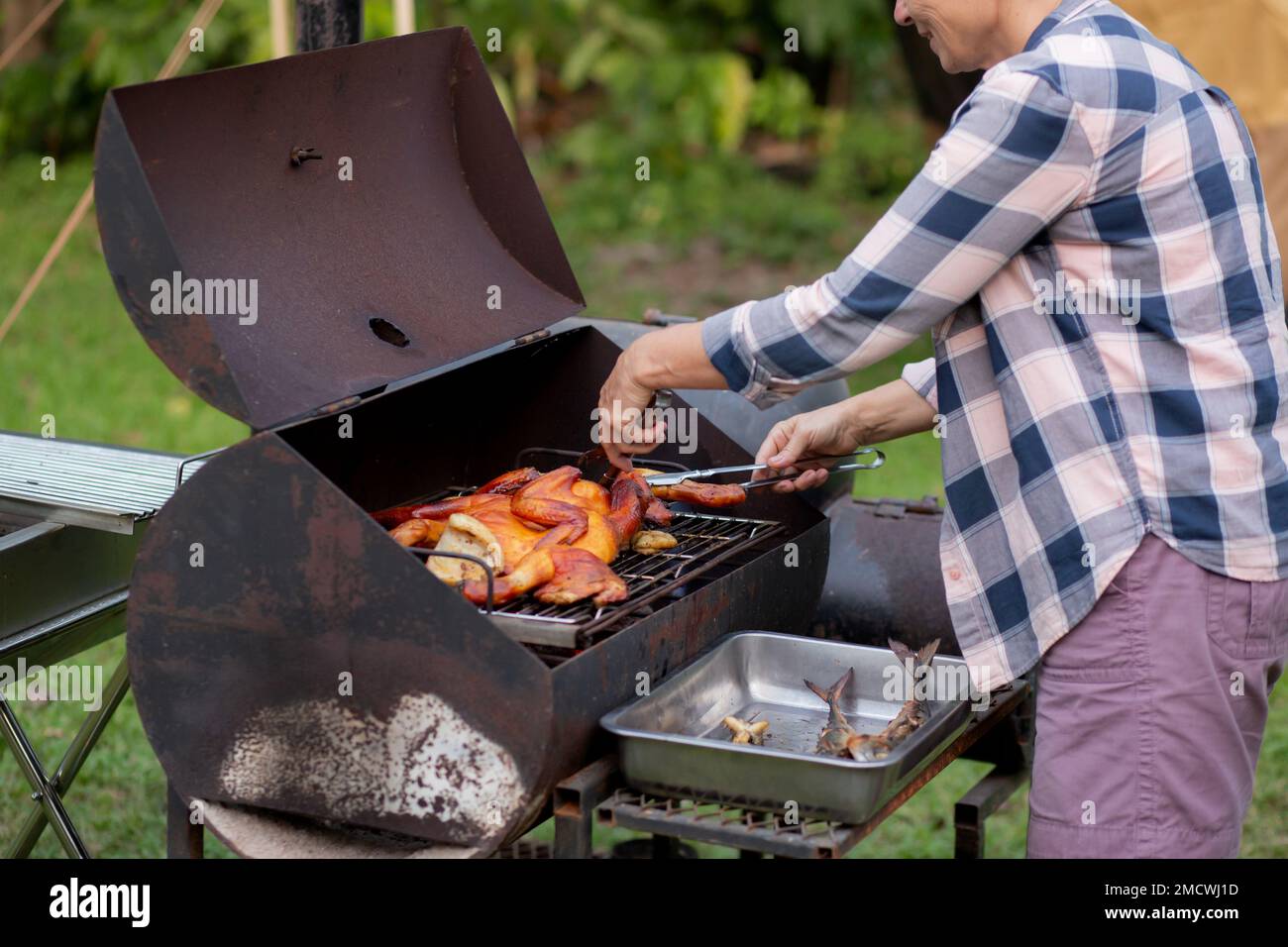 Seniorin grillt Hühnerfleisch für die Zubereitung des Essens im Camping, Senioren machen Freizeitaktivitäten und Freizeit-Picknick, während sie am Wochenende Fleisch braten, Stockfoto