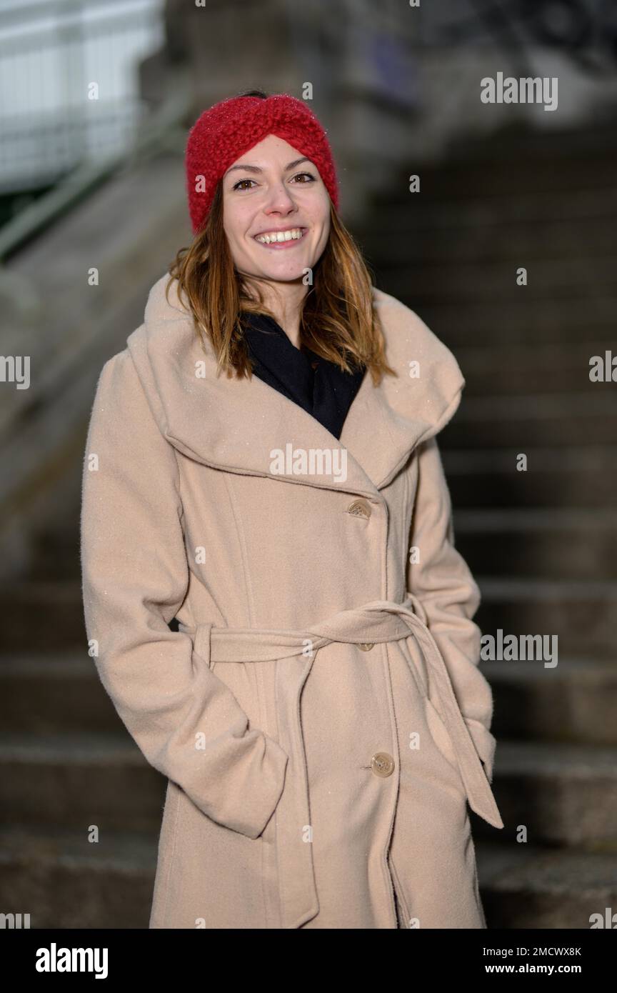 Lächelnde junge Frau mit langen braunen Haaren, einem hellbraunen Stoffmantel und einer roten Wollmütze auf einer Treppe aus Natursteinen Stockfoto