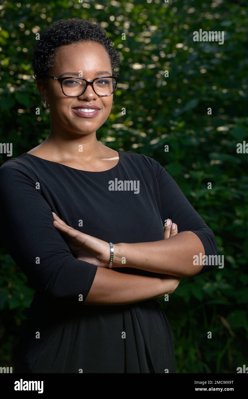 Eine ziemlich selbstbewusste junge afroamerikanische Frau mit kurzen Haaren, einer großen Brille in einem schwarzen Kleid vor einem grünen Busch im Sonnenlicht Stockfoto