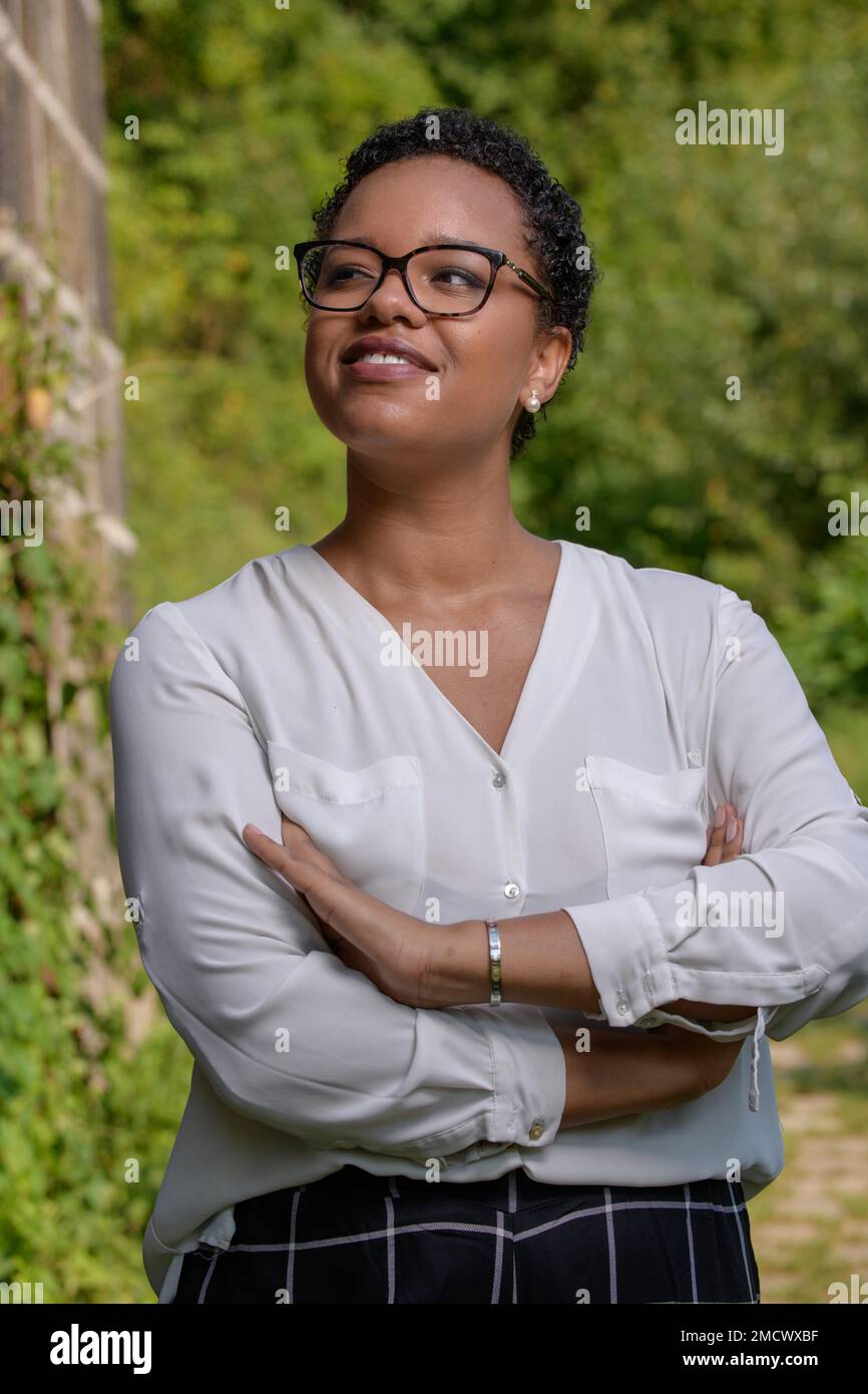 Eine ziemlich selbstbewusste junge afroamerikanische Frau mit kurzen Haaren, einer großen Brille in einer weißen Bluse vor einem grünen Busch im Sonnenlicht Stockfoto