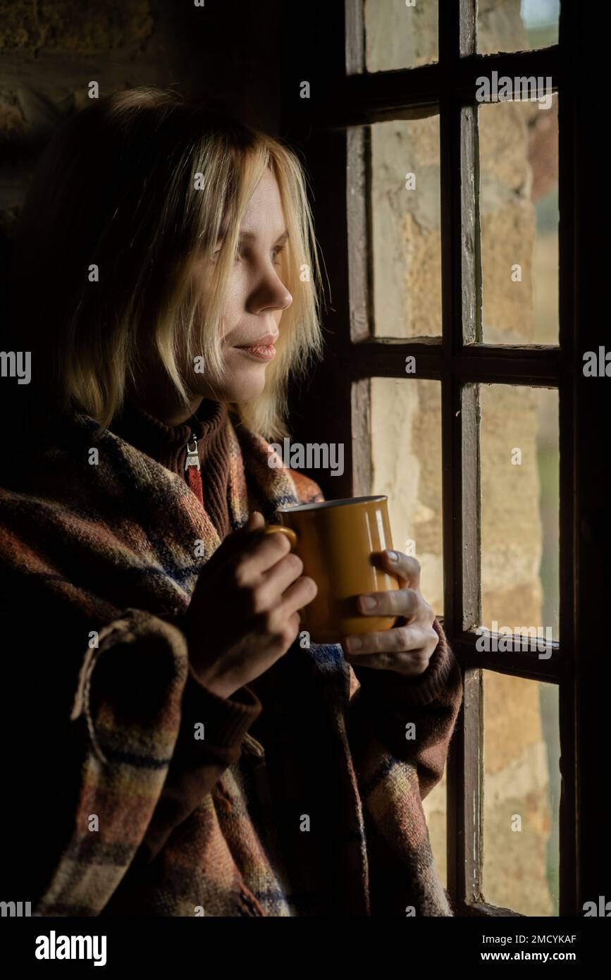 Eine Frau trinkt Kaffee im Dunkeln am Fenster. Eine Frau in ihren Vierzigern. Nachdenklichkeit, Nachdenklichkeit. Stockfoto
