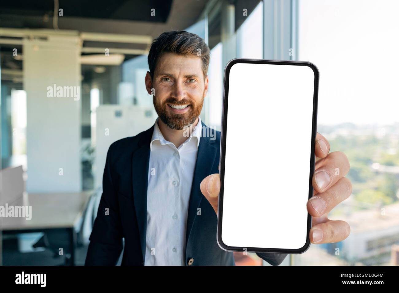 Glücklicher Geschäftsmann mittleren Alters, der im Büro ein Smartphone mit weißem Bildschirm zeigt, ein Modell für die mobile App oder das Websitedesign Stockfoto