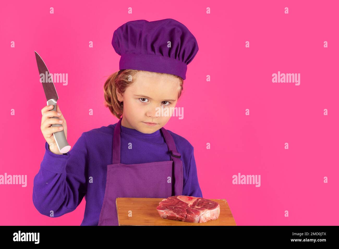 Kinderkoch hält Schneidebrett mit Fleischsteak und Messer. Kinderkoch, Studioporträt. Kinder kochen. Ein Junge mit Schürze und Kochmütze. Stockfoto