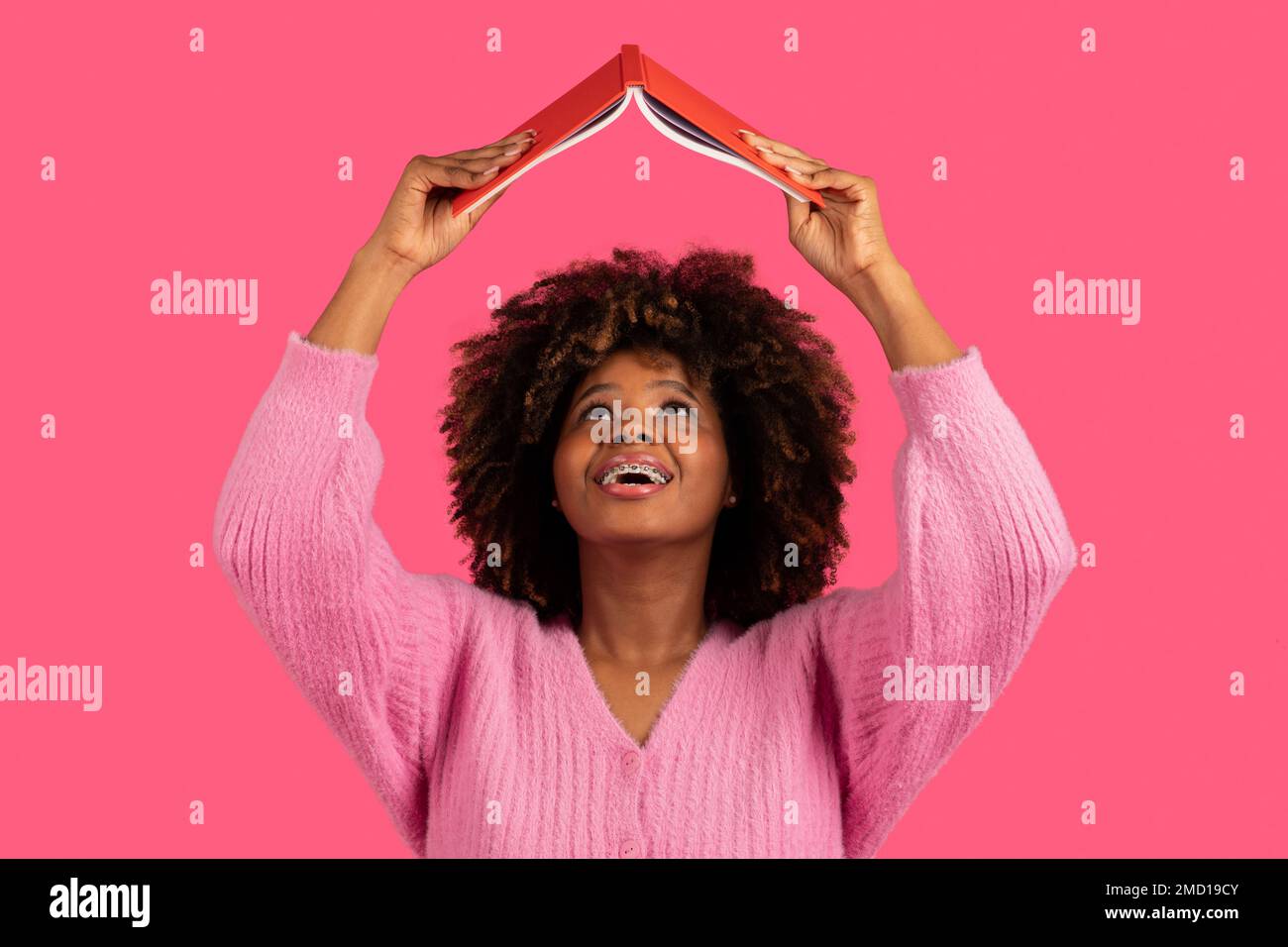 Fröhliche, lustige, junge afroamerikanische, lockige Studentin mit Zahnspangen hebt die Bücher hoch und liest, sucht Informationen Stockfoto