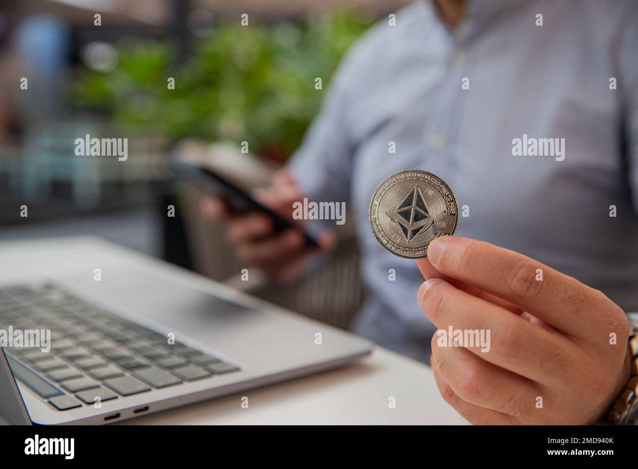 Ethereum-Münze in der Hand eines Händlers, der den Markt mit seinem Laptop und Smartphone überwacht Stockfoto