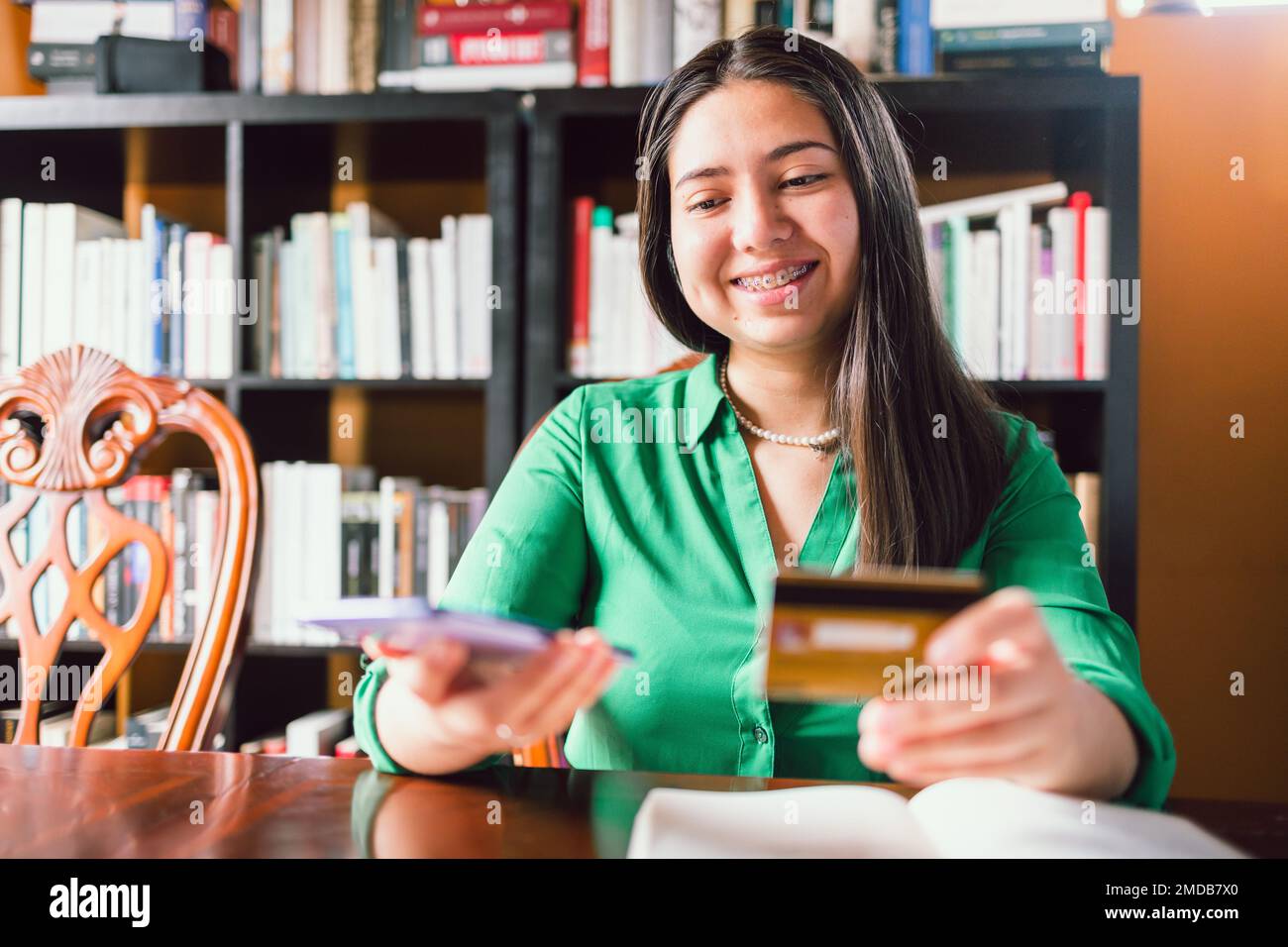 Lächelnde junge Frau, die eine Kreditkarte benutzt, um online in ihrer persönlichen Bibliothek zu kaufen. Online-Shopping Stockfoto