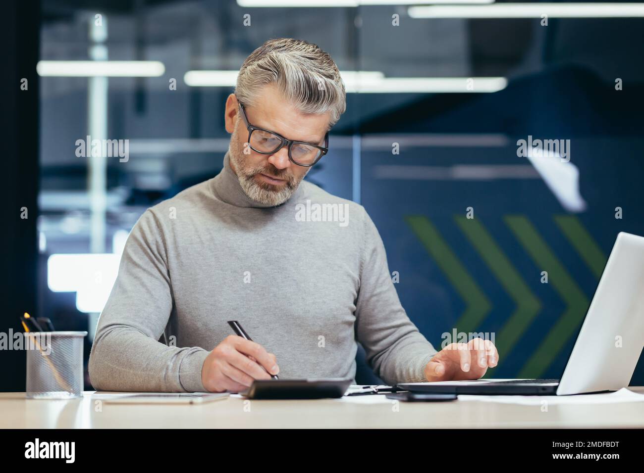 Ein seriöser Geschäftsmann im Büro, der mit einem Laptop arbeitet, ein grauhaariger Senior-Mann, der am Schreibtisch sitzt. Stockfoto