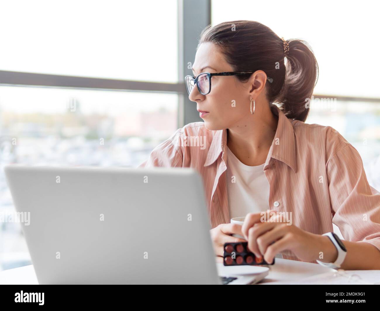 Die Stirnrunzeln runzelnde Frau schaut auf Medizinpillen, während sie mit einem Laptop arbeitet. Psychische Probleme, emotionaler Burnout oder Kopfschmerzen. Modernes Büro bei Co-working c Stockfoto
