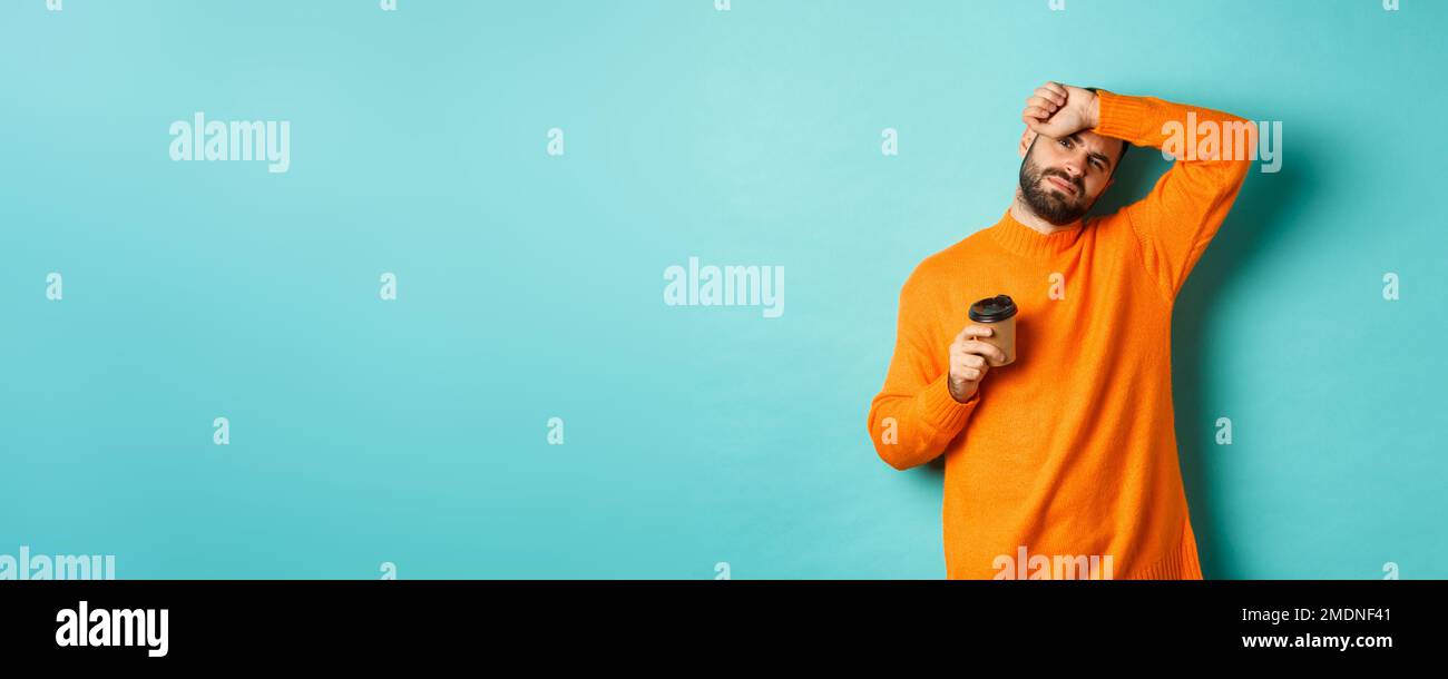 Müde Erwachsene Mann Pause von der Arbeit und Kaffee trinken, wischen Schweiß von der Stirn und sieht erschöpft, in orange Pullover über türkis stehen Stockfoto