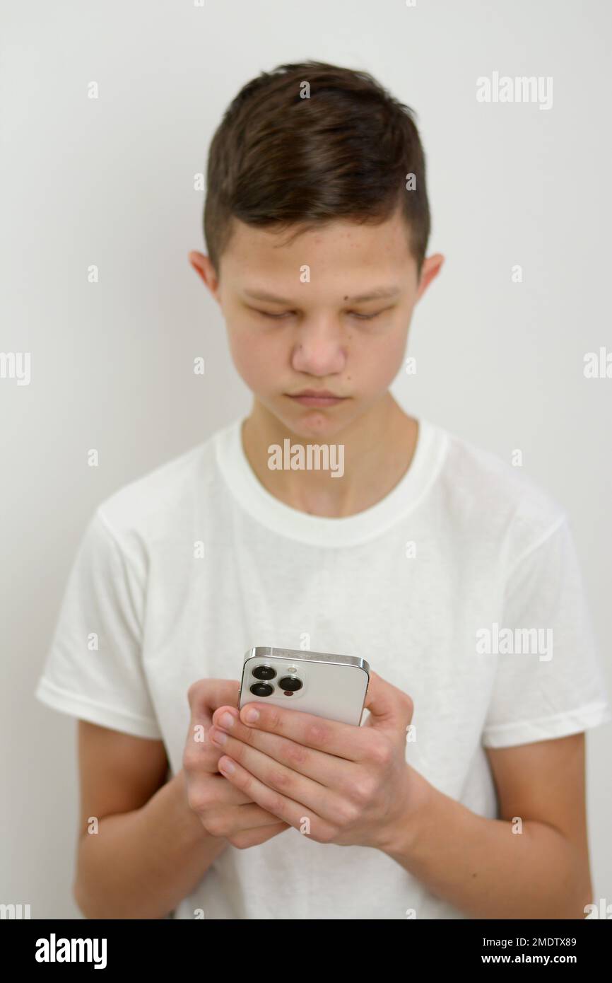 Ein 10-15-jähriger Junge steht auf weißem Hintergrund mit Telefon in den Händen. Er spielt Spiele oder surft in sozialen Netzwerken. Sein Gesicht ist fokussiert und schaut auf die Smartphone-Ohren herunter, kleines Pfotenohr, schwarzes Haar Stockfoto