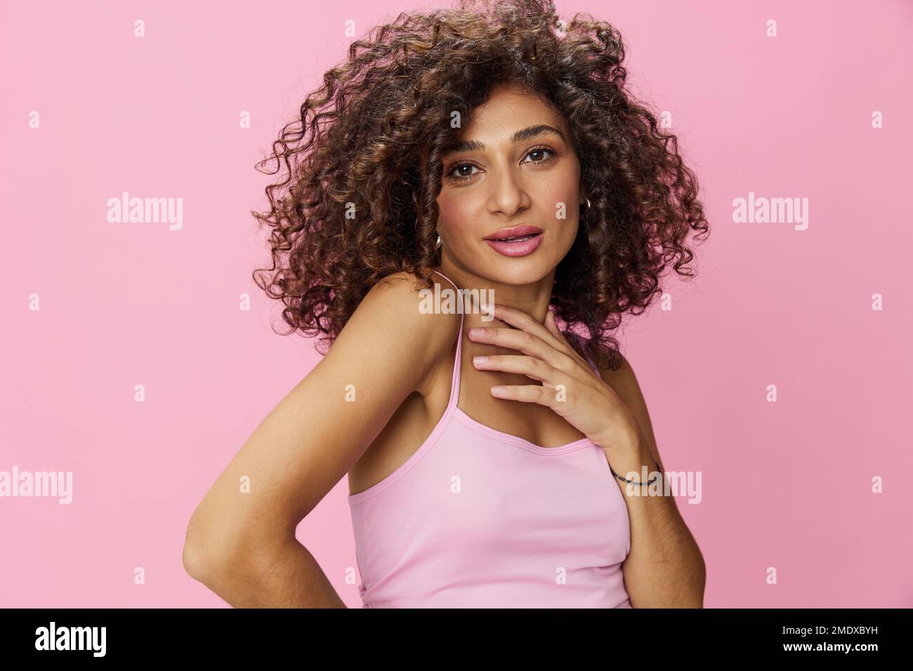 Eine Frau mit lockigem Afro-Haarmodell posiert auf pinkfarbenem Hintergrund in einem pinkfarbenen T-Shirt, bietet Bewegungsfreiheit und Tanz, schaut in die Kamera, lächelt mit Zähnen und Stockfoto