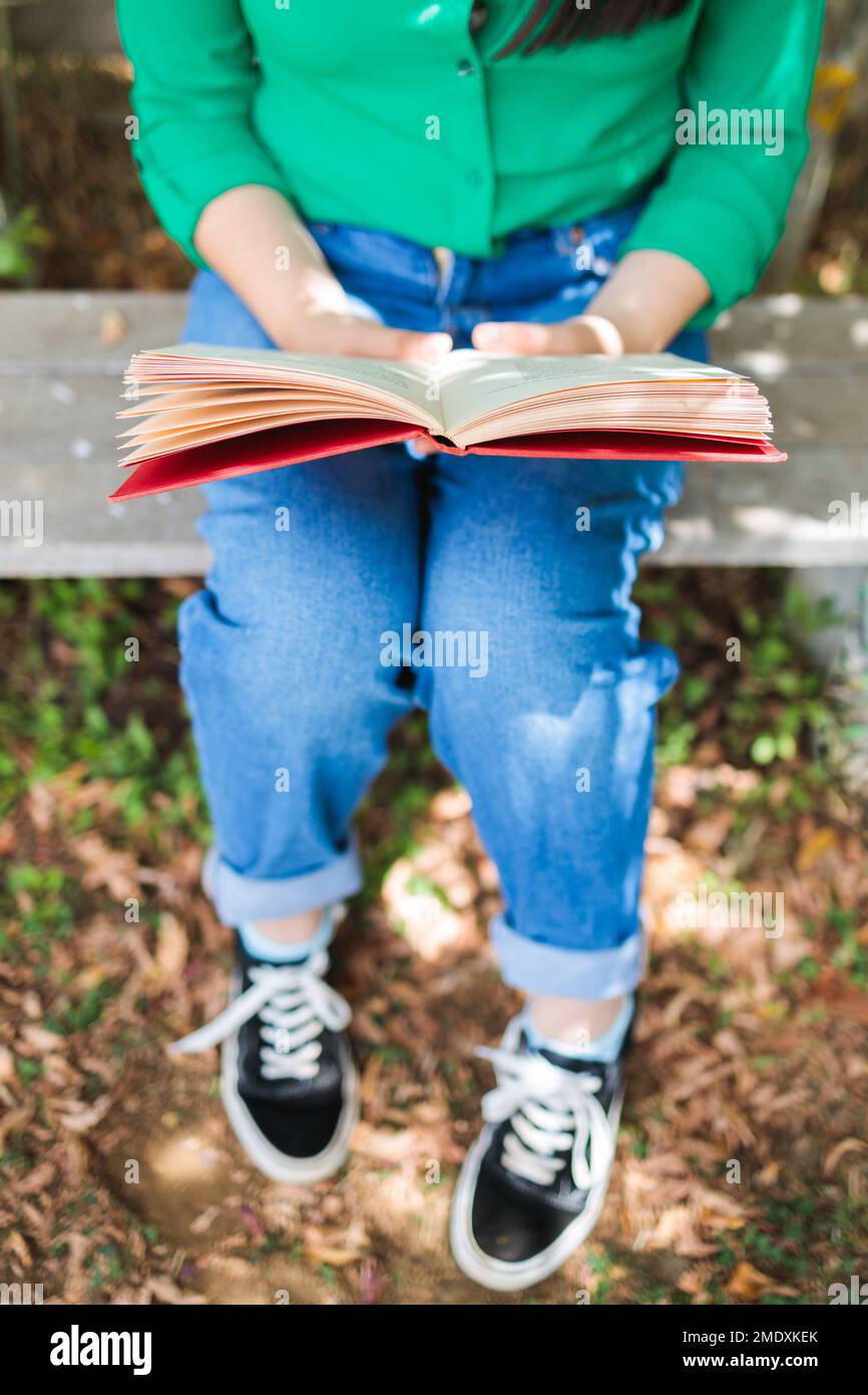 Ein unbekanntes Mädchen mit einer grünen Bluse und Jeans, das im Garten ein Buch mit Sonnenlicht liest. Weltbuchtag Stockfoto