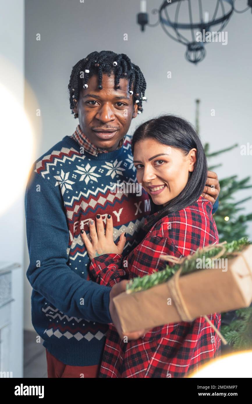 Ein vielseitiges junges Paar, das sich mit Geschenken in der Nähe des Weihnachtsbaums umarmt. Hochwertiges Foto Stockfoto