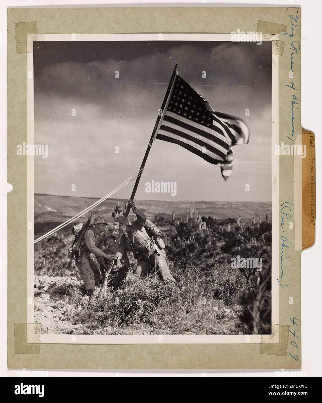 Die Flaggenheber von Okinawa. Die Marines und Küstenwachmänner imitieren die jetzt berühmte Flagge, die auf Iwo Jimas vulkanischem Berg Suribachi gehisst wird, und beugen sich dem Anpflanzen der ersten Sterne und Streifen, die über Okinawa winken. Die Flagge fing die Brise auf einer Höhe, von der aus das Meer auf beiden Seiten zu sehen war. Das viel gelobte Heraufsetzen der Fahne von Iwo Jima hat Soldaten zu einer heftigen Rivalität inspiriert, bei jeder neuen Inseleroberung mitzuwirken, um die „ersten“ Farben zu entfalten. Diese Flagge wurde gespendet von der Crew einer von der Küstenwache bemannten LST, die Männer und Material auf Okinawas Sand entladete. Stockfoto