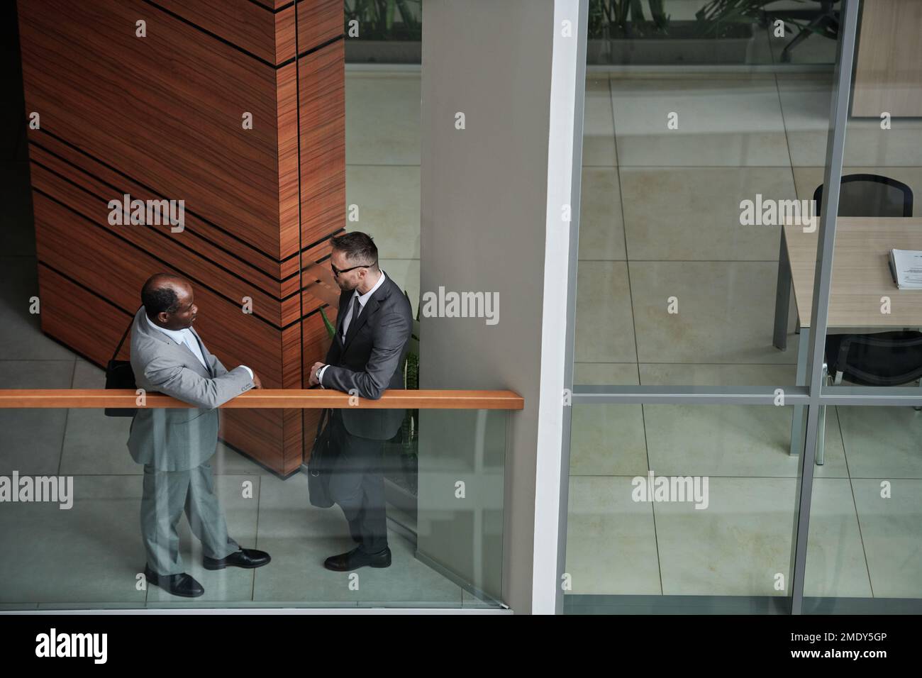 Teil der Inneneinrichtung des modernen Businesscenters mit zwei interkulturellen männlichen Angestellten in formeller Kleidung, die sich beim Treffen am Geländer unterhalten Stockfoto