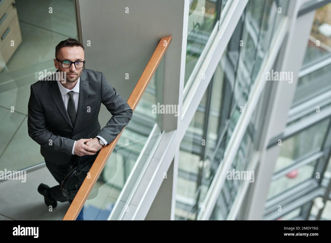 Junger, selbstbewusster Chief Executive Officer in formeller Kleidung, steht am durchsichtigen Geländer im Büro und schaut in die Kamera Stockfoto