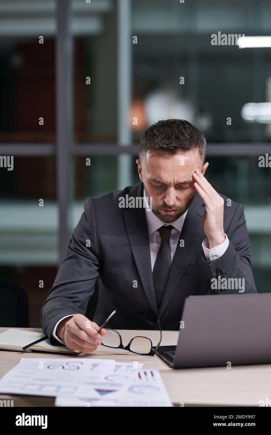 Überarbeiteter und müder Geschäftsmann in formeller Kleidung, der am Schreibtisch sitzt, sich über den Laptop bückt und den Tempel berührt, während er versucht, sich zu konzentrieren Stockfoto