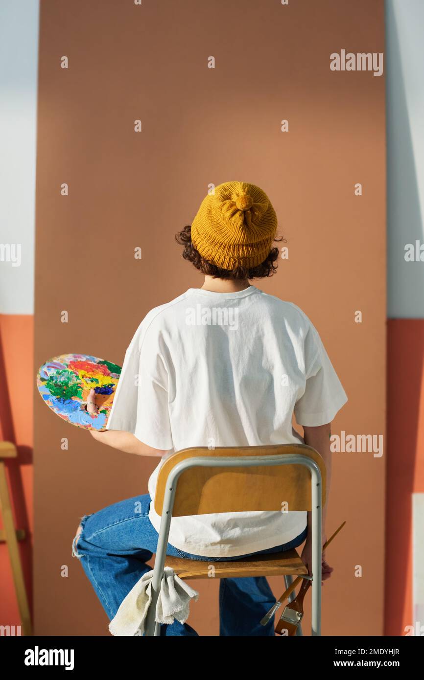Rückansicht einer jungen Künstlerin in Freizeitkleidung, die vor einem blanken Werkstück sitzt und für die Malerei zukünftiger Meisterwerke vorbereitet ist Stockfoto