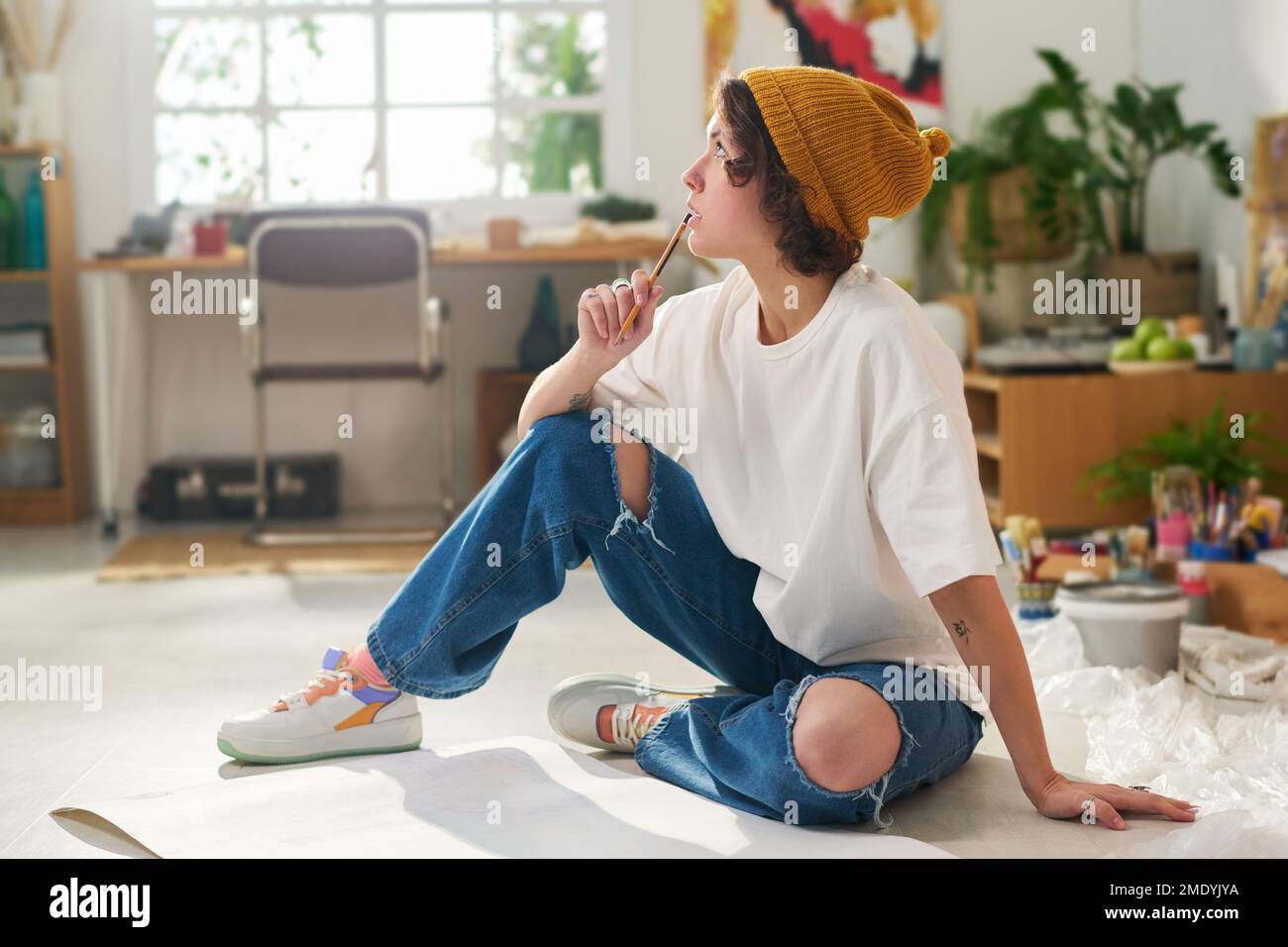 Eine junge nachdenkliche Frau in Freizeitkleidung, die einen Bleistift am Mund hält, während sie mit Papier auf dem Boden sitzt und an neue Kunstwerke denkt Stockfoto