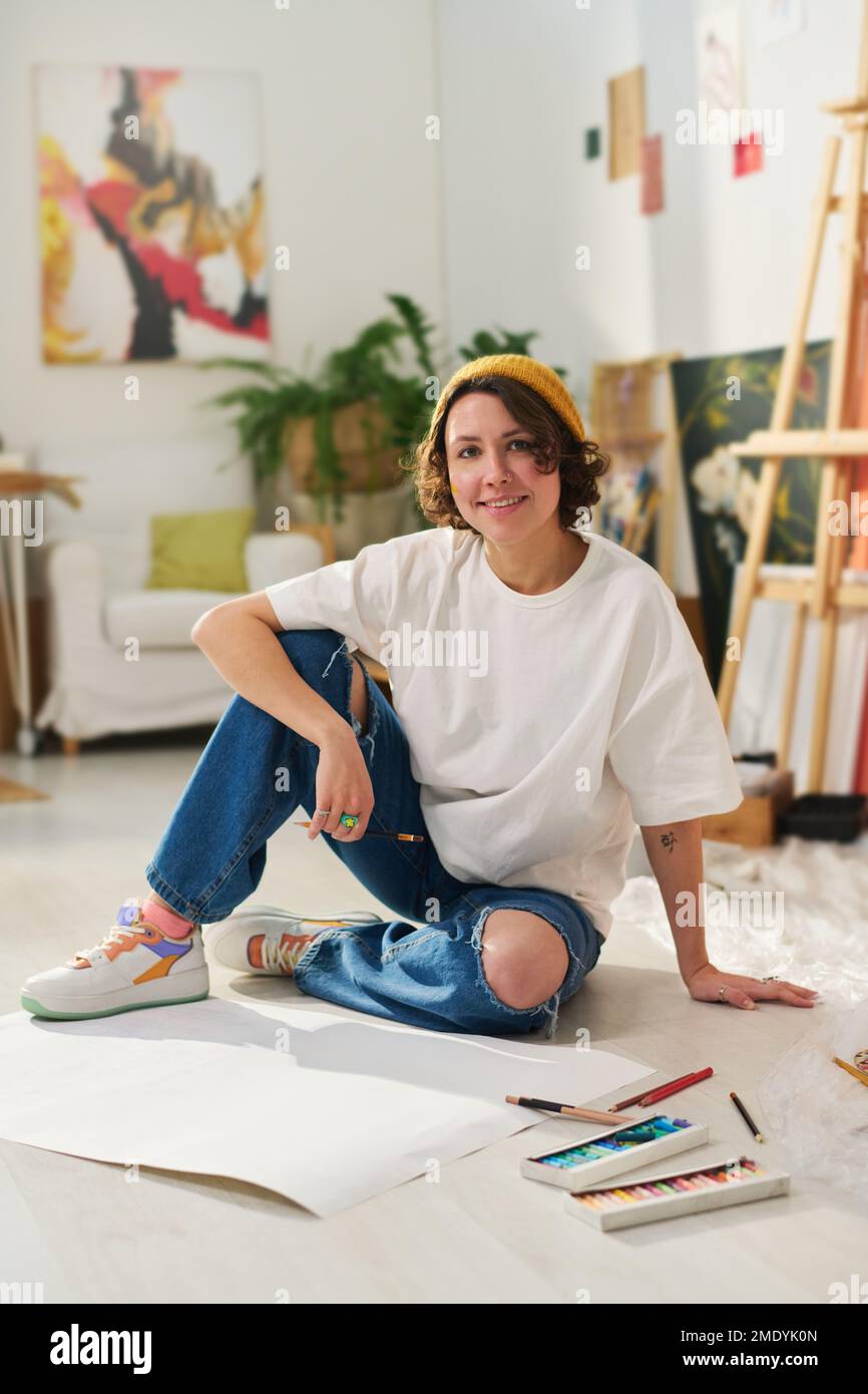 Junge lächelnde Künstlerin in Freizeitkleidung, die auf dem Boden des Heimstudios sitzt und beim Erstellen neuer Skizzen in die Kamera schaut Stockfoto