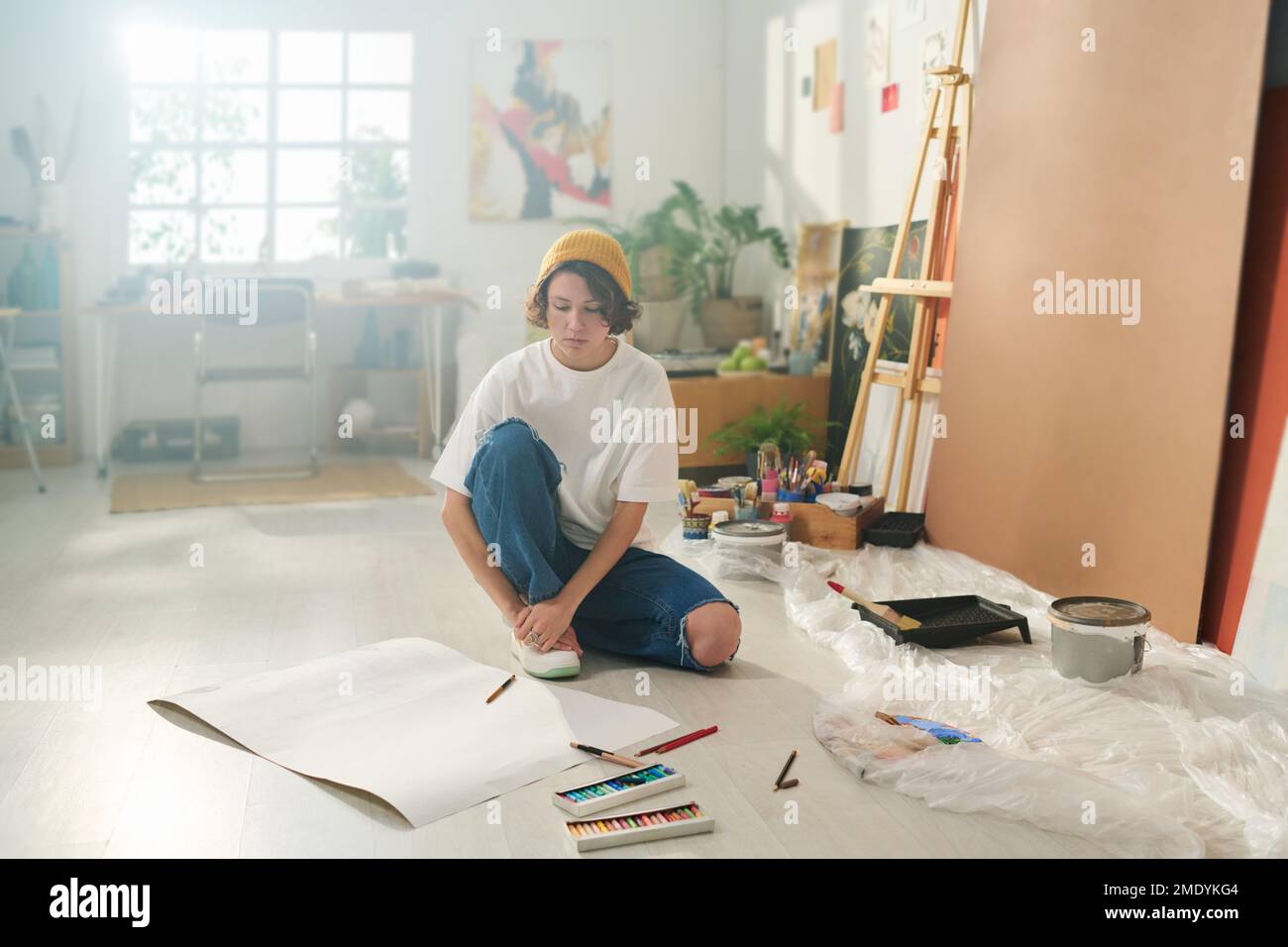 Junger Künstler in Freizeitkleidung sitzt auf dem Boden des Heimstudios und sieht sich leeres Papier an, während er sich für die Gestaltung neuer Kunstwerke inspirieren lässt Stockfoto