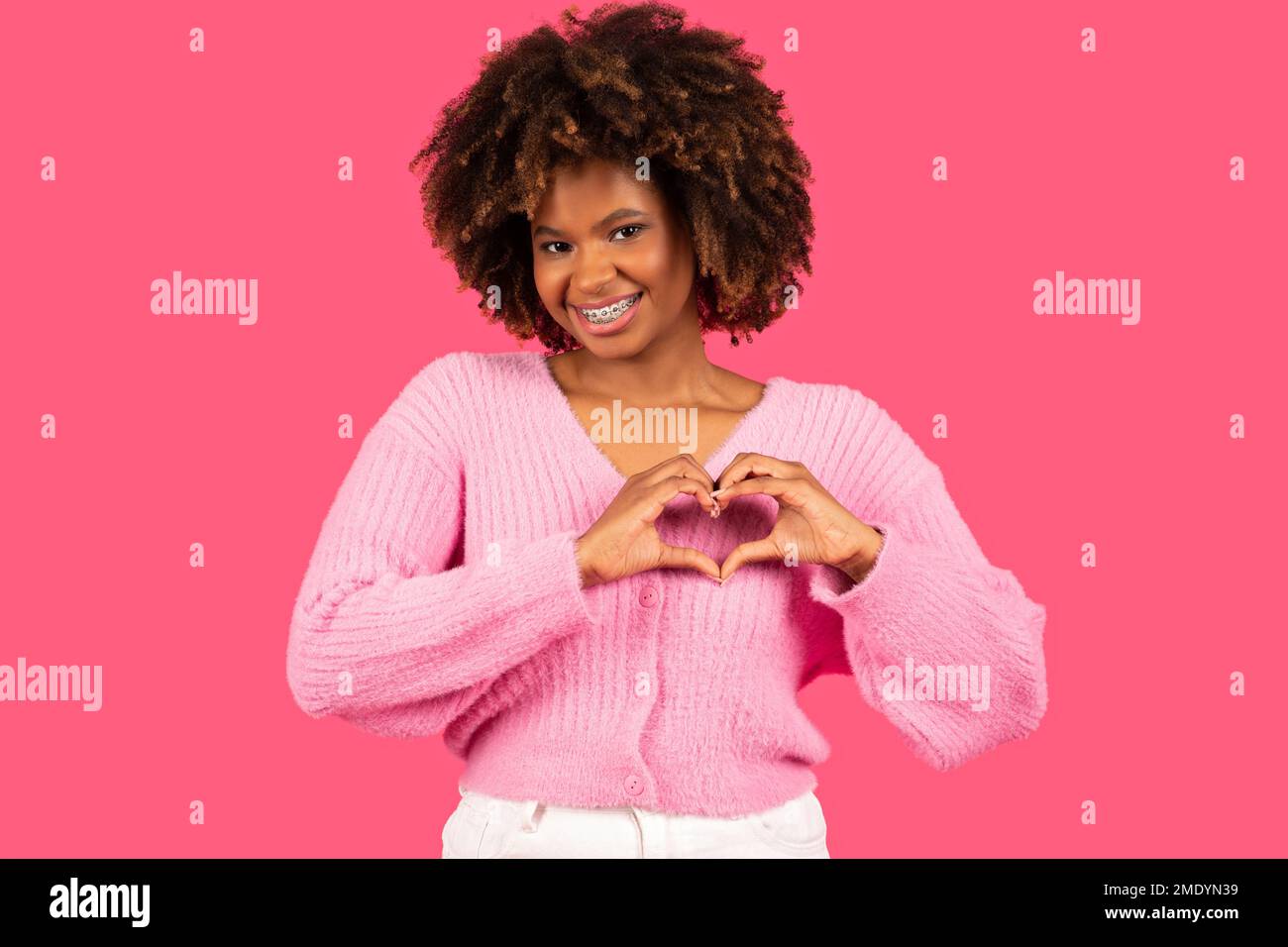 Glückliche, hübsche, junge, afroamerikanische, lockige Frau mit Zahnspangen in lässigem Outfit, mit Herzzeichen und Händen Stockfoto