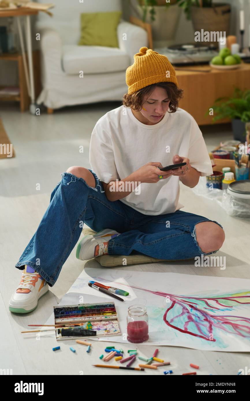 Junge kreative Malerin mit Smartphone fotografiert neue Kunstwerke, die sie auf Papier gemalt hat, während sie auf dem Boden saß Stockfoto