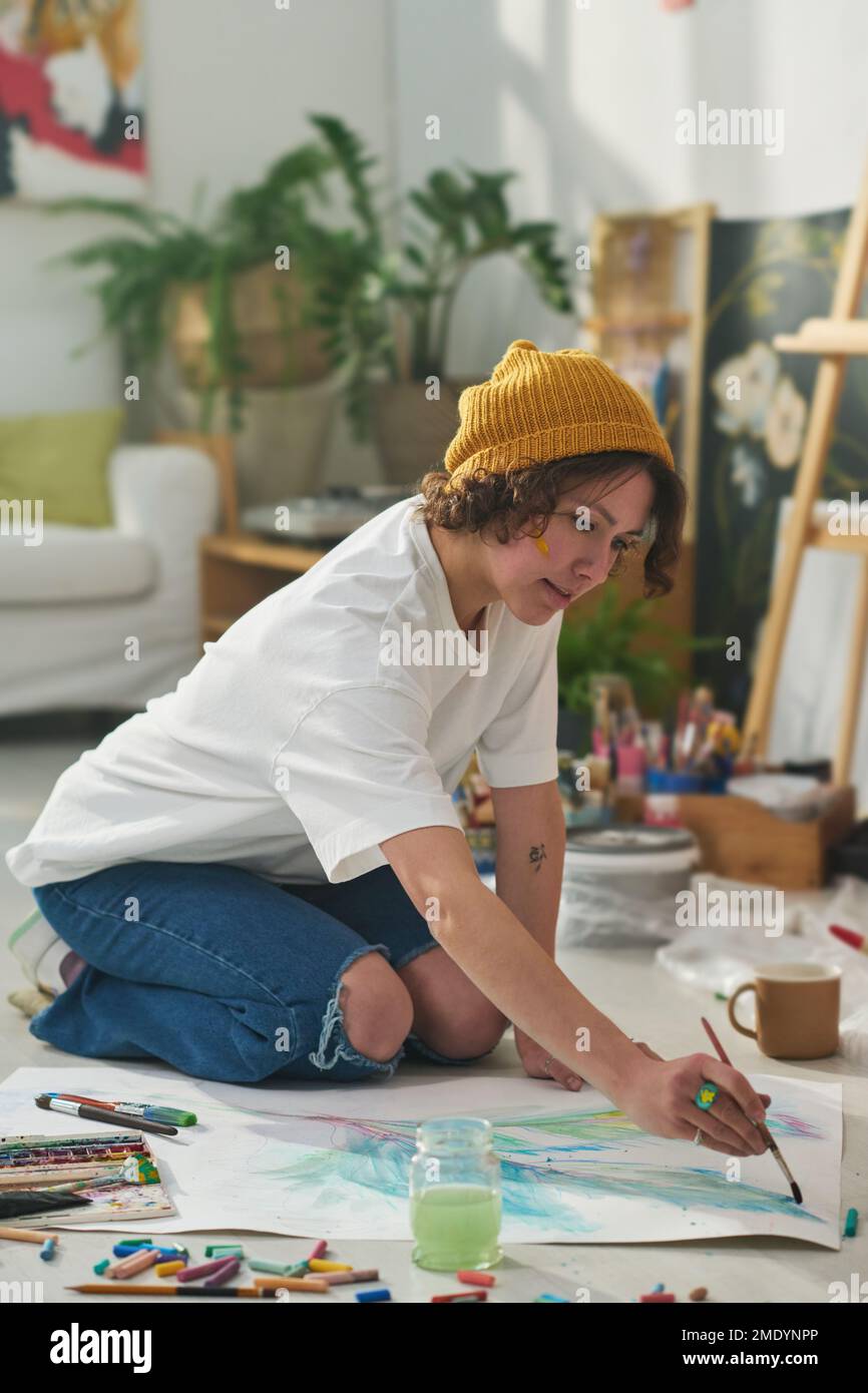 Junge kreative Frau mit Pinsel- und Wasserfarben, die auf dem Boden des Studios oder der Werkstatt malen, während sie neue Kunstwerke kreiert Stockfoto