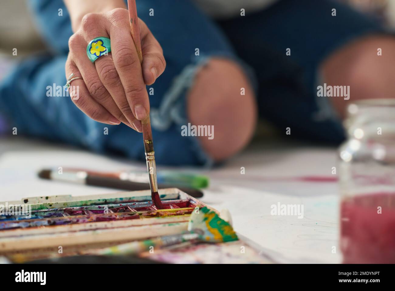 Konzentrieren Sie sich auf die Hand einer jungen kreativen Frau mit dem Bijou-Ring, der den Pinsel in Aquarellfarben legt, während er auf dem Boden der Werkstatt sitzt und Kunstwerke kreiert Stockfoto