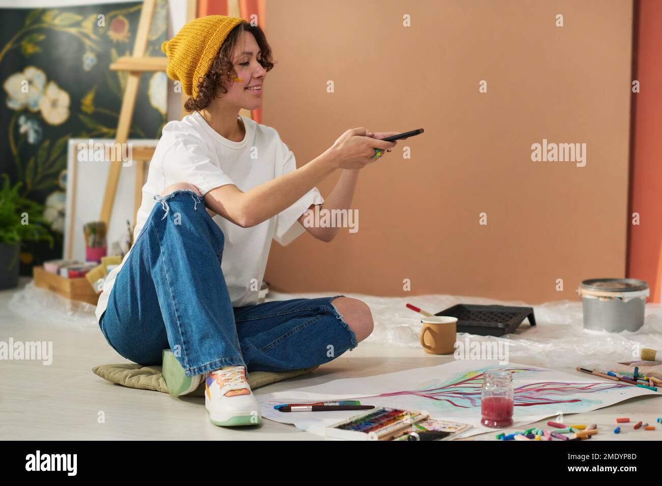 Fröhliche junge kreative Malerin mit Smartphone auf dem Boden und Foto von neuen Kunstwerken, die mit Aquarellen auf Papier gemalt wurden Stockfoto