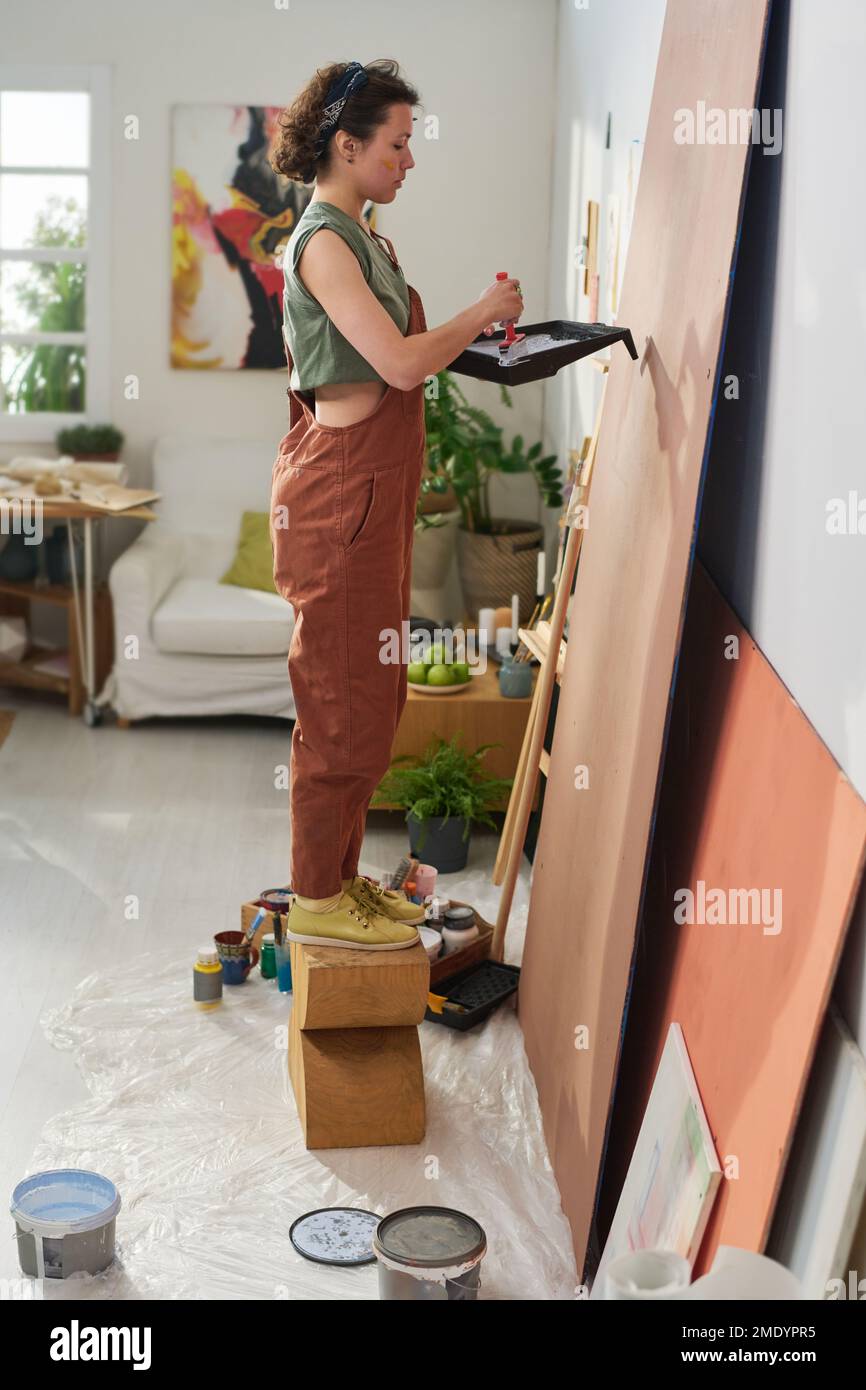 Seitenansicht einer jungen kreativen Malerin in Freizeitkleidung, die ein neues Meisterwerk kreiert, während sie im Heimstudio vor dem Panel steht Stockfoto
