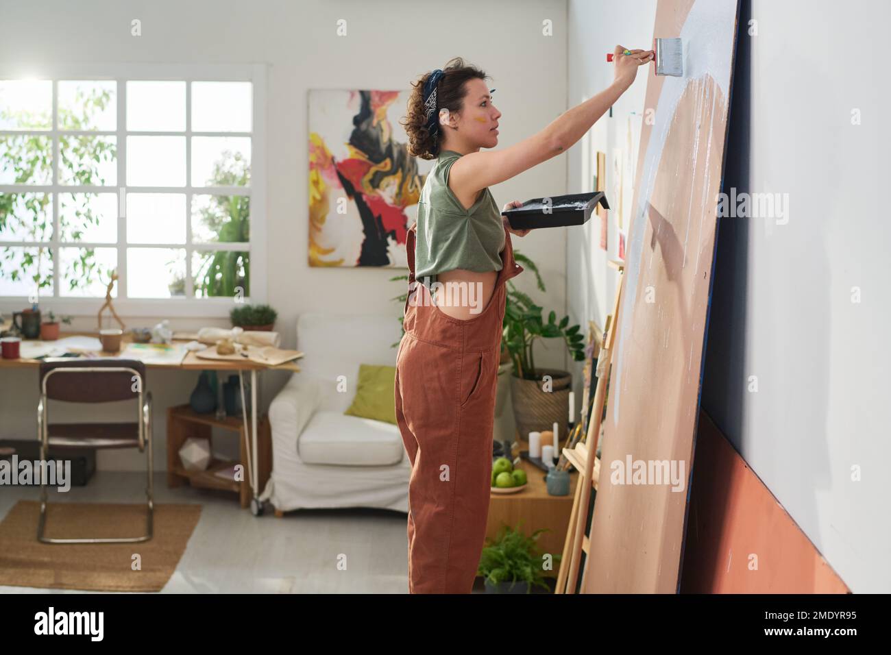 Seitenansicht eines jungen Künstlers in Freizeitkleidung, der ein Tablett mit Farbe und Pinsel hält und ein neues abstraktes Meisterwerk auf der Platte kreiert Stockfoto