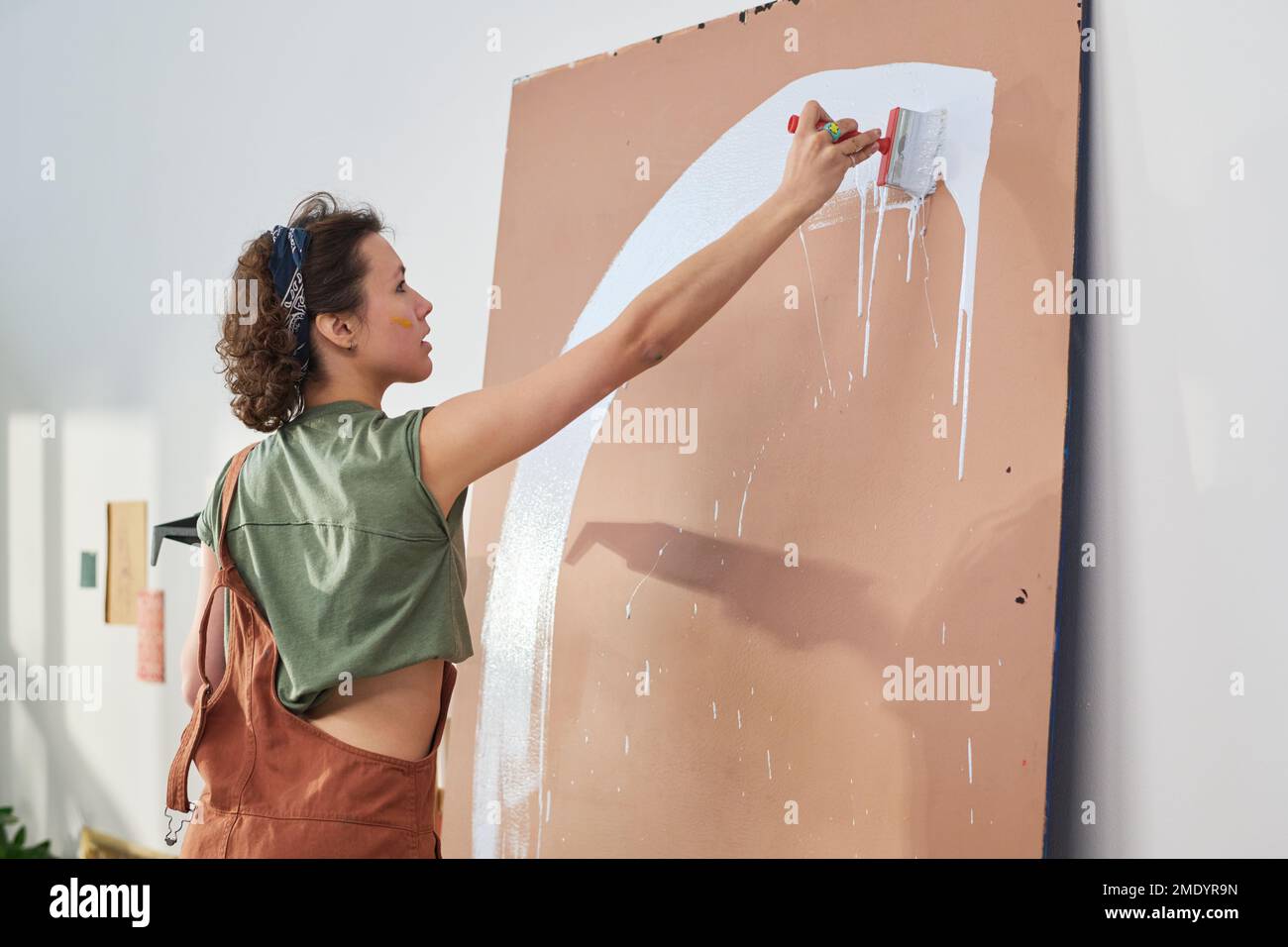 Eine junge, inspirierte Kunsthandwerkerin in Freizeitkleidung, die Pinsel und weiße Farbe verwendet und neue Kunstwerke auf dem Bedienfeld vor der Kamera kreiert Stockfoto