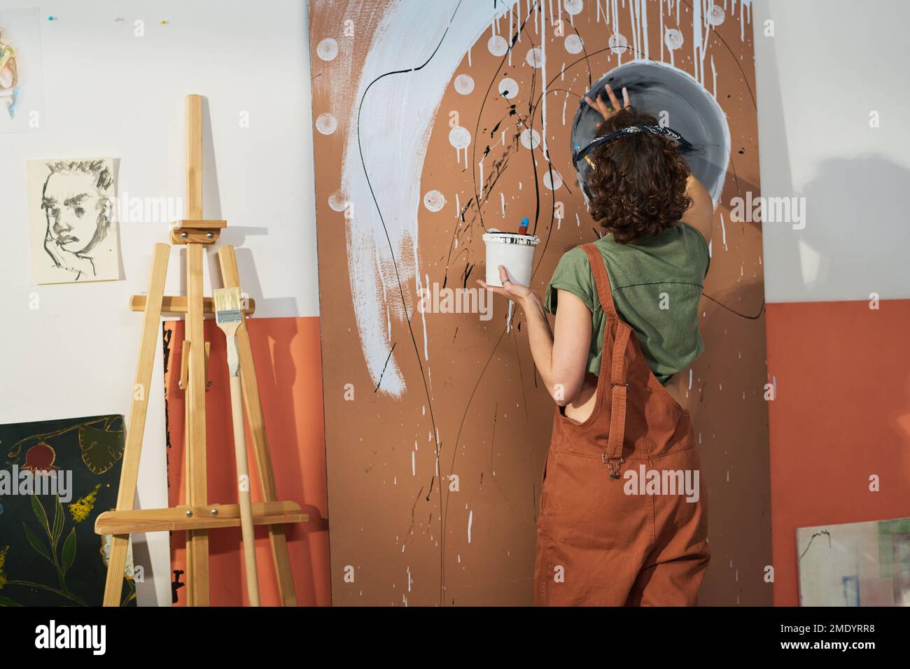 Rückansicht einer jungen kreativen Frau mit Farben, die neue abstrakte Kunstwerke kreieren, während sie in ihrem Heimstudio vor dem Panel steht Stockfoto