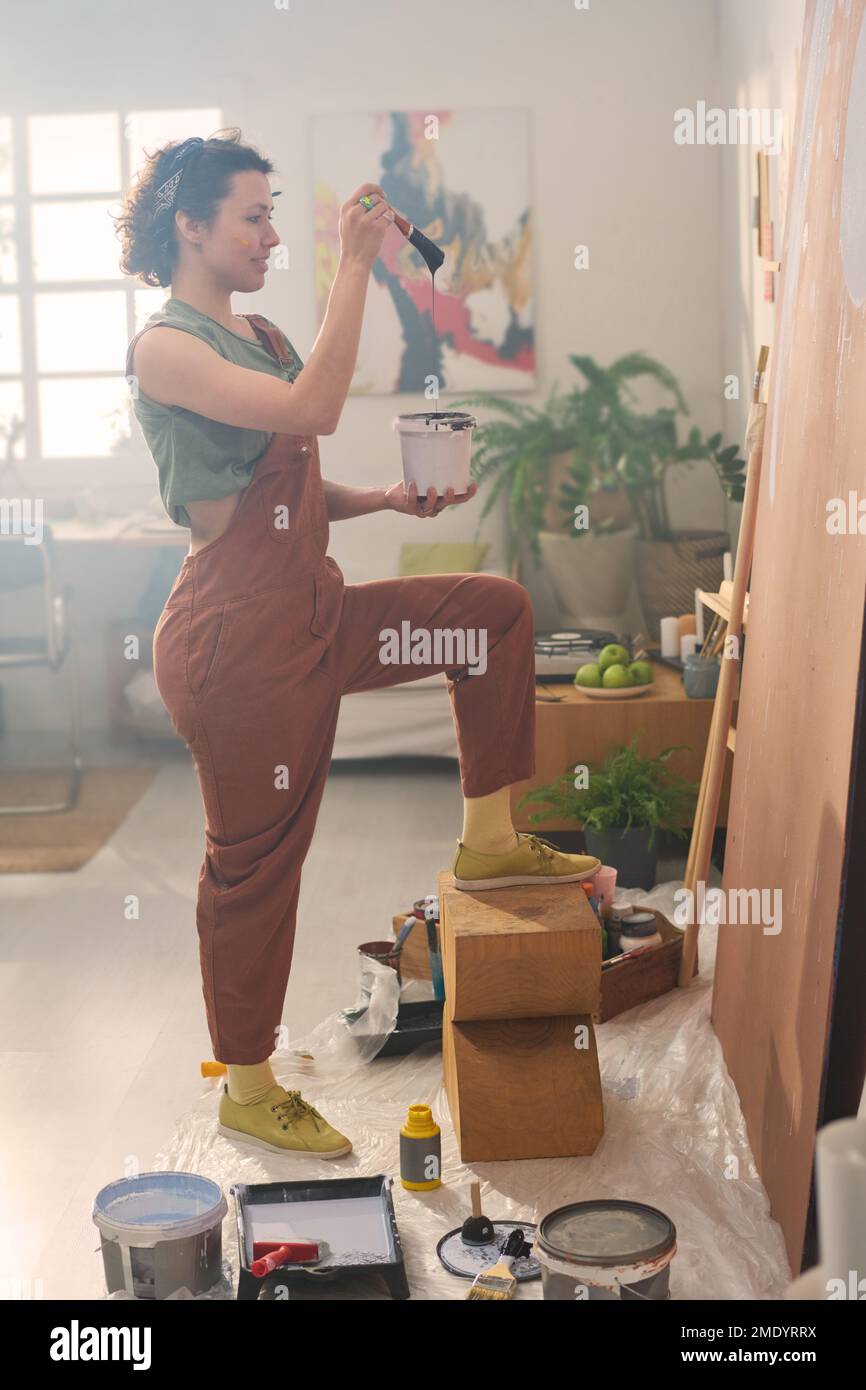 Seitenansicht einer jungen braunen Künstlerin mit kleinem Eimer und Pinsel, die vor einem großen Panel steht und ein neues Meisterwerk kreiert Stockfoto