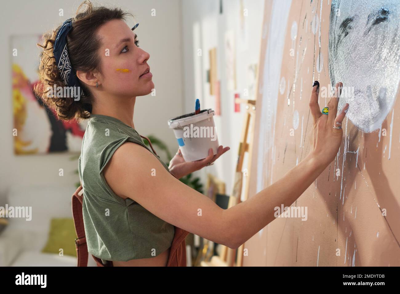 Ein junges kreatives Mädchen mit einem kleinen Eimer Farbe, das ein neues Meisterwerk kreiert, mit den Händen an einer großen Wand ihrer Werkstatt oder ihres Heimstudios Stockfoto