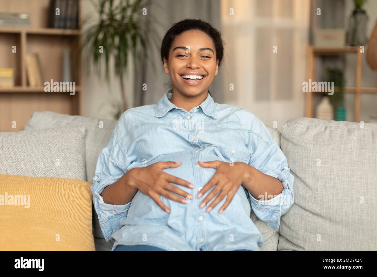 Schwangerschaftsfreude. Glückliche afroamerikanische schwangere Frau, die den Bauch umarmt und vor der Kamera lächelt, zu Hause auf dem Sofa sitzt Stockfoto