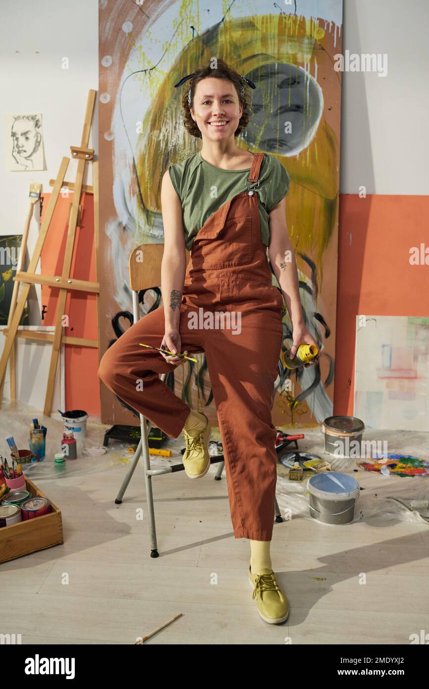 Junge lächelnde Künstlerin oder Maler im Overall sitzt im Zentrum des Studios gegen eine Leinwand mit Meisterwerk und schaut in die Kamera Stockfoto
