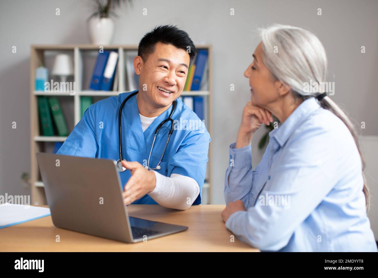 Glücklich glücklich gut aussehender koreanischer Arzt mittleren Alters zeigt Laptop einer alten Kaukasierin in der Klinik Stockfoto