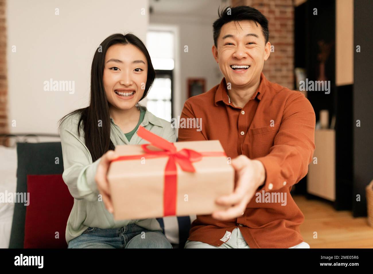 Glückliches asiatisches Paar, das sich in einer Geschenkbox in der Kamera ausstreckt und lächelt, zu Hause auf der Couch sitzt Stockfoto
